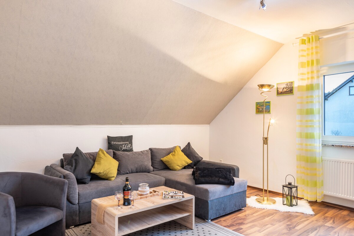 A spacious living room features a large couch with decorative pillows, an elegant lighting fixture, and a coffee table. Large windows allow natural light to brighten the space, while a cozy corner seating arrangement invites relaxation.