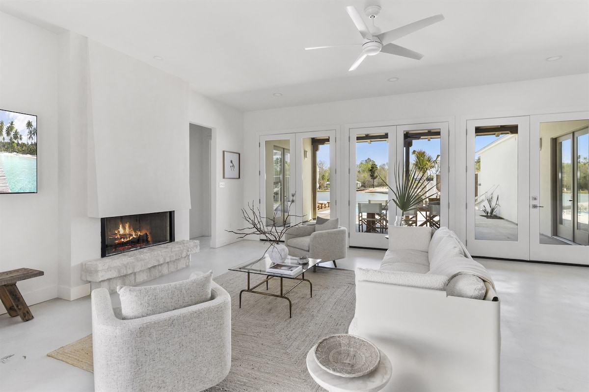 An airy living area features a cozy fireplace set in a minimalist white wall. Two plush armchairs and a sectional sofa are arranged around a simple round coffee table. Large glass doors open to the outdoor space, allowing natural light to illuminate the room.