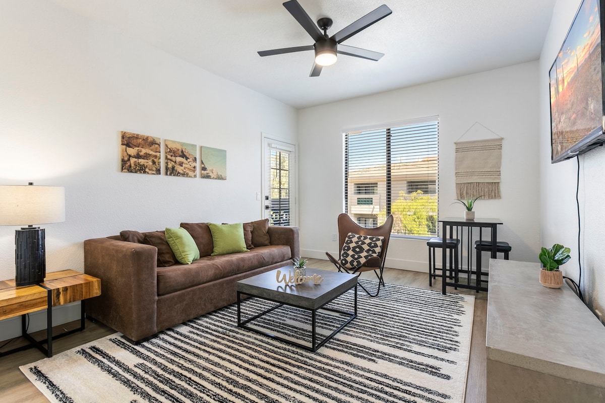 A light-filled living area features a brown sofa adorned with green accent pillows. A minimalist coffee table sits on a striped rug, complemented by a stylish chair. A flat-screen TV is mounted on the wall, and large windows provide views of the surrounding landscape.