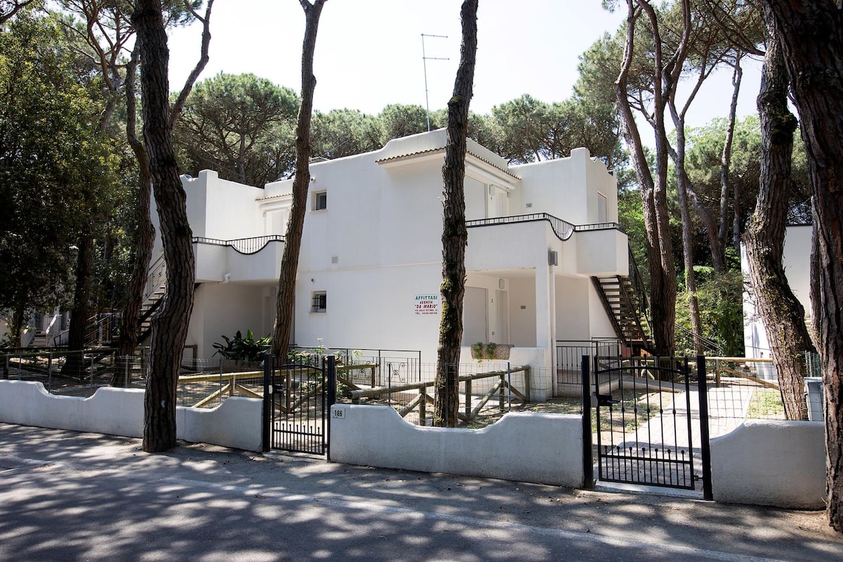 The two-story villa is set amidst tall pine trees, featuring a white exterior that contrasts with the greenery. A gated entrance provides access, with a staircase leading to the upper level visible on the right. The surrounding area is tranquil and natural.