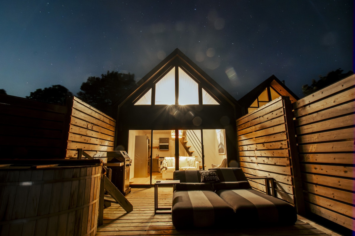 A modern loft retreat is illuminated under a starry night sky, revealing an inviting deck. A spacious hot tub is positioned nearby, along with a sleek outdoor seating area. The warm glow from interior windows hints at a cozy atmosphere within.