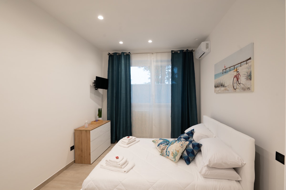 A bright and spacious bedroom is featured, with a comfortable bed adorned with fluffy pillows. A dresser is positioned against one wall, alongside a mounted television. Light filters through sheer curtains, and artwork depicting a bicycle by the sea adds a subtle decorative touch.