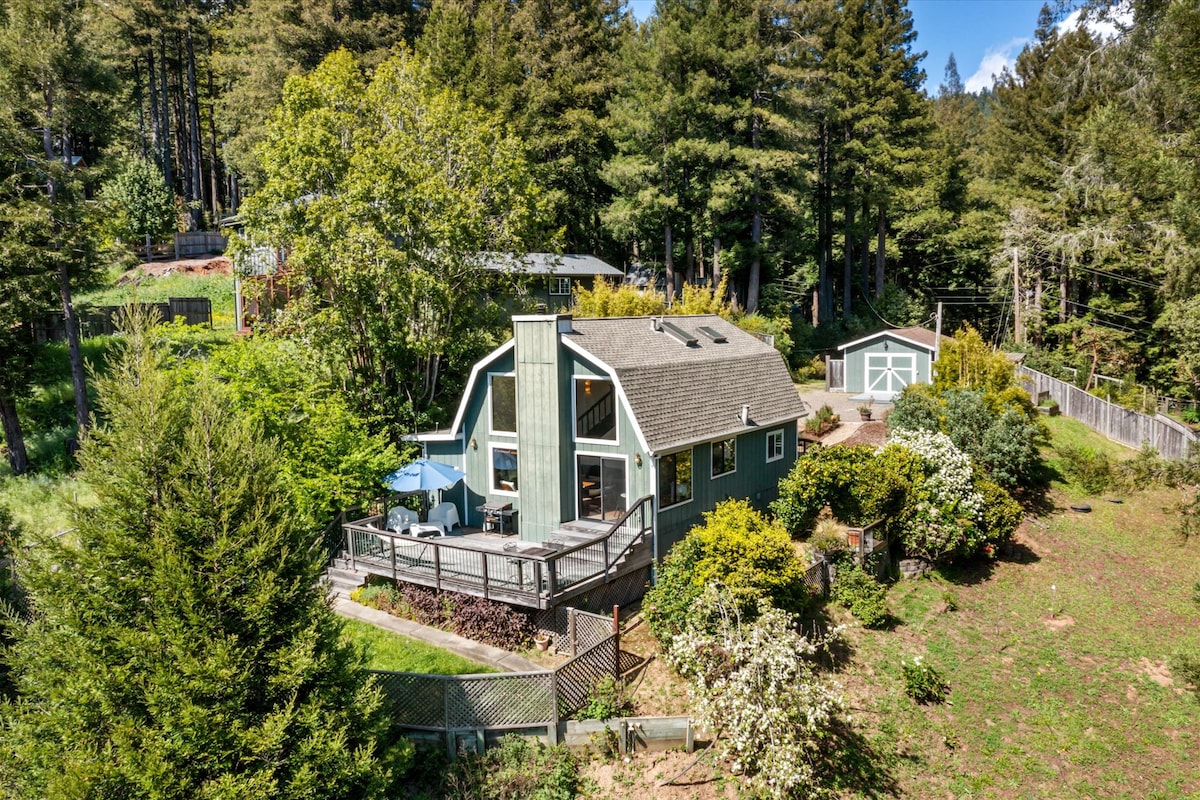 The exterior of the home is shown, featuring a spacious deck surrounded by greenery and flowers. Large windows allow natural light to fill the interior. The structure is set within a forested area, providing a serene and private ambiance.