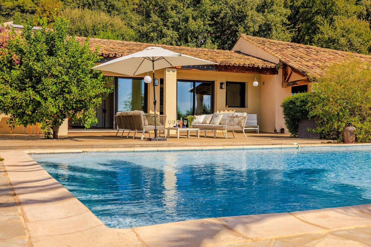 A tranquil outdoor space features a clear swimming pool with sun loungers arranged around it. An umbrella provides shade for a seating area, which is positioned near large glass doors opening into the villa. Lush greenery surrounds the property, enhancing the serene atmosphere.