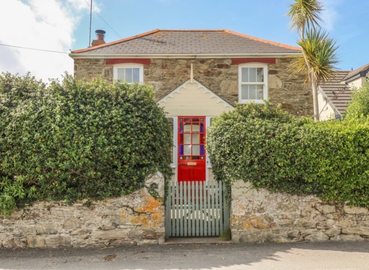 A charming cottage exterior is framed by lush greenery and a low stone wall. The entrance features a vibrant red door, complemented by white window frames. A wooden gate opens into a welcoming pathway, bordered by dense hedges and accompanied by a palm tree in the foreground.