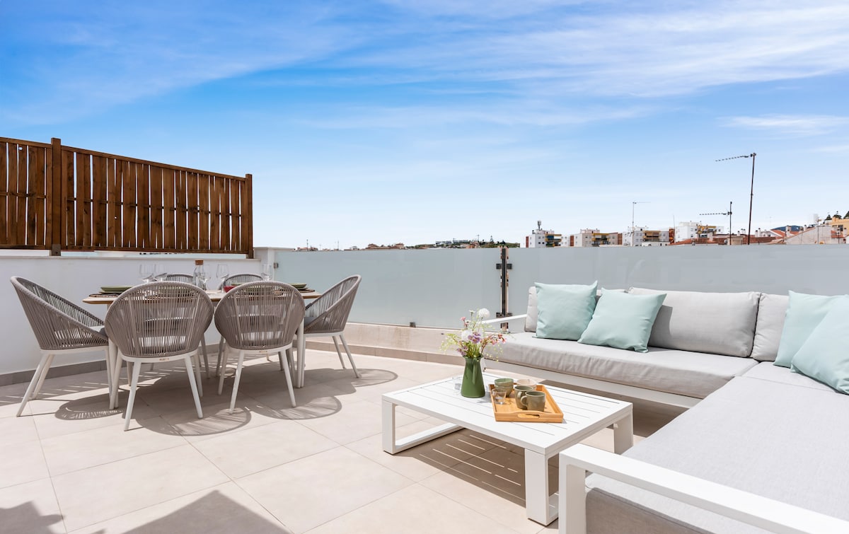 An outdoor terrace is presented, featuring a lounge area with a sectional sofa and a coffee table. A dining table set for four, accompanied by comfortable chairs, is also visible. The backdrop showcases a clear sky and views of Estepona's architectural landscape.
