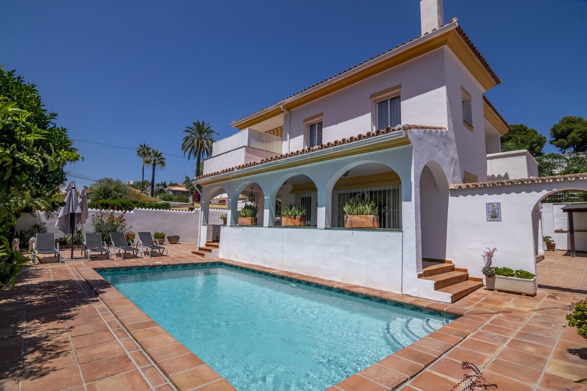 The villa features an inviting swimming pool surrounded by a tiled terrace. Arched openings provide a glimpse of the exterior's greenery. The two-story structure is complemented by a clear blue sky and palm trees, enhancing the outdoor space's bright and airy feel.