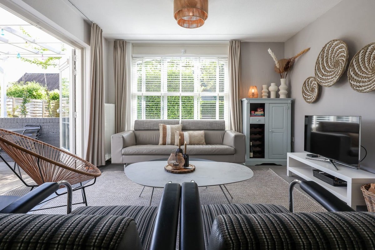 A welcoming living space features a comfortable sofa arranged next to two stylish armchairs. A round coffee table takes center stage, complemented by decorative details on the walls and a flat-screen TV. Natural light filters in through large windows, creating an airy atmosphere.
