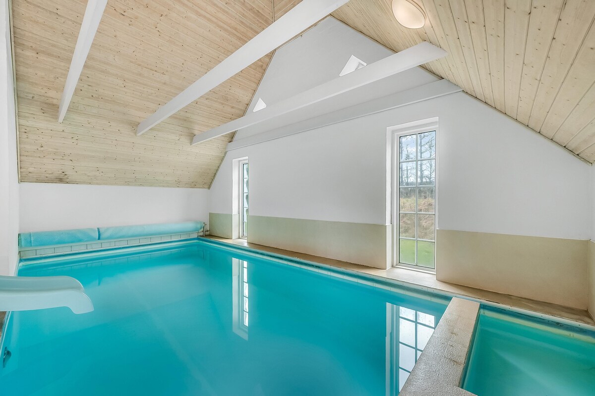 An indoor swimming pool is featured with a vibrant blue surface, inviting for relaxation and fun. High ceilings are adorned with wooden beams, and large windows allow natural light to fill the space. A water slide is visible at one end, enhancing the playful atmosphere.