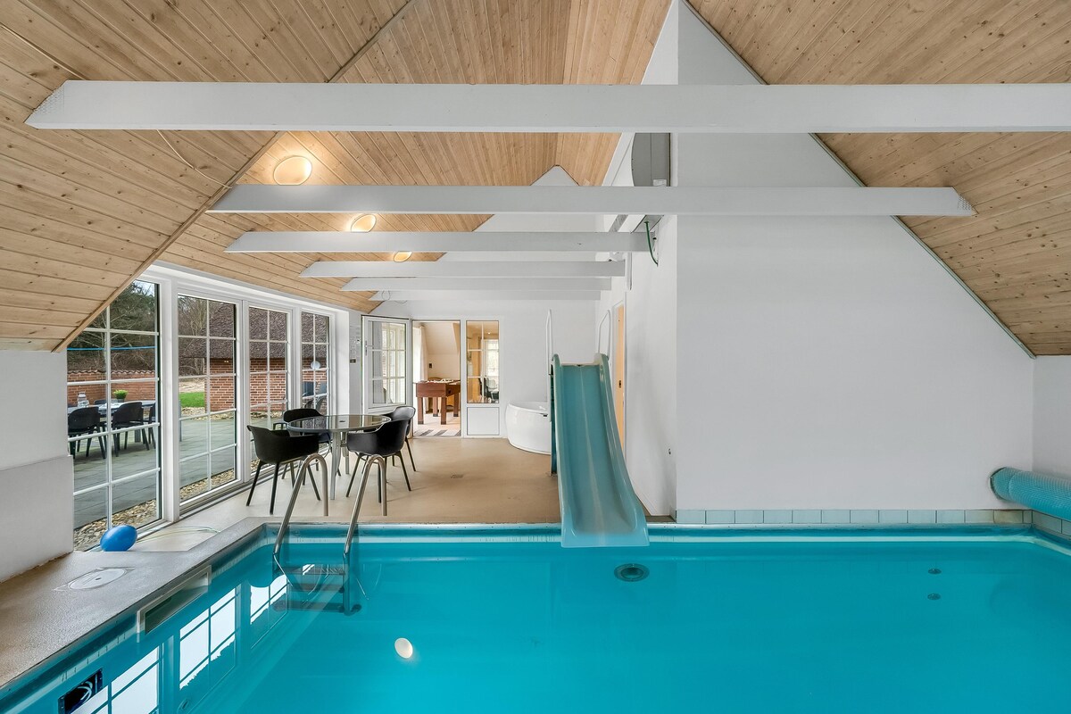 An indoor swimming pool features crystal-clear water complemented by a fun water slide. Natural light filters through large glass doors, illuminating the space. A comfortable dining area with black chairs is positioned nearby, providing a relaxed atmosphere for guests.
