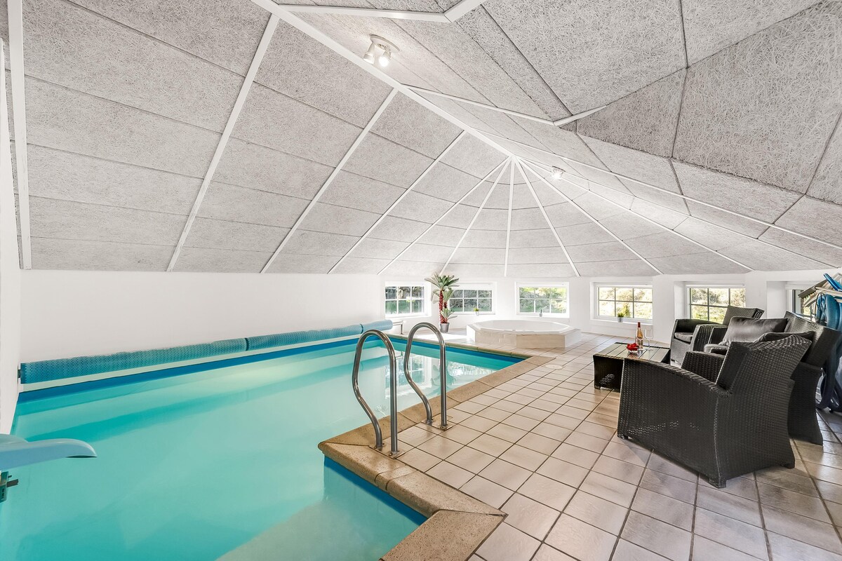 An indoor swimming pool area is showcased, featuring a smooth, blue pool adjacent to a water slide. The room includes comfortable seating with dark wicker chairs and a textured ceiling that enhances the bright atmosphere. Large windows allow natural light to fill the space.