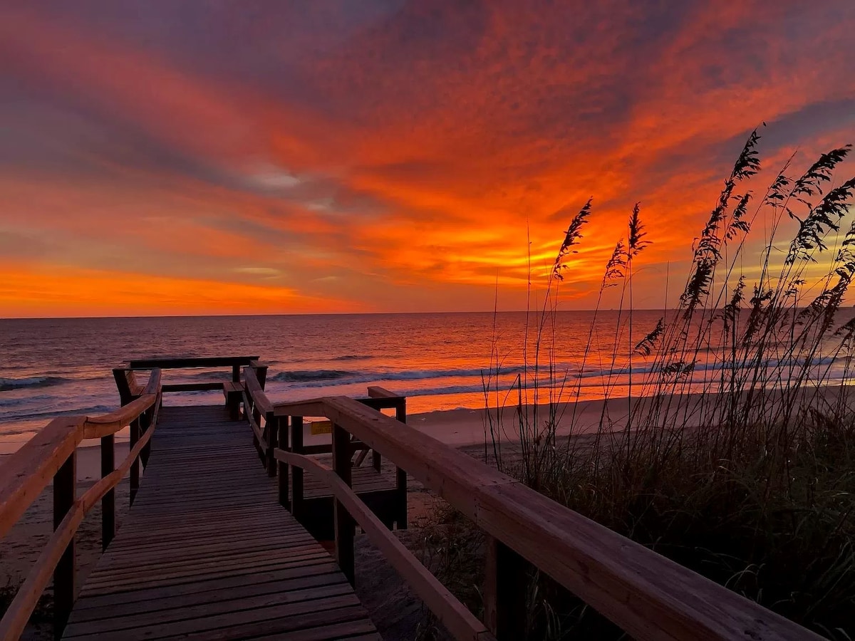 A wooden boardwalk leads toward the beach, framed by tall grasses. The vibrant sunset paints the sky in shades of orange, pink, and purple, reflecting on the calm ocean waves. The scene captures a serene coastal moment.