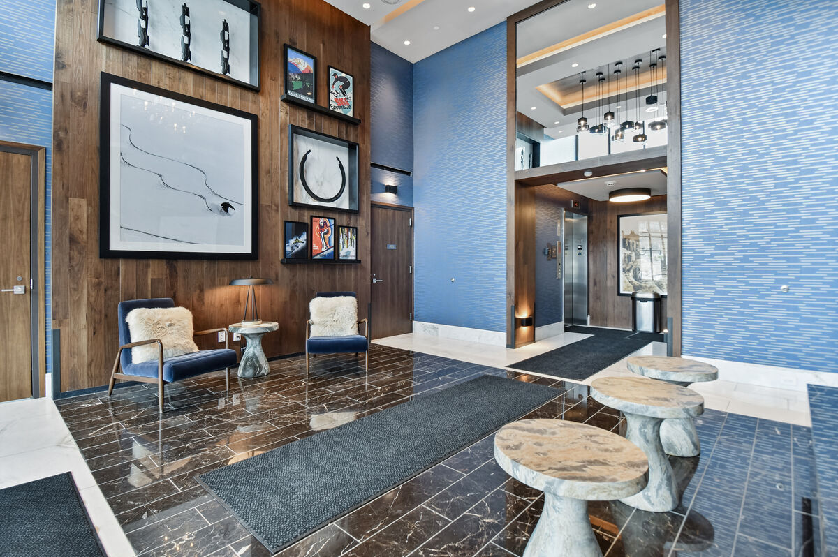 The lobby features modern design elements with blue textured walls and dark marble flooring. Stylish seating areas incorporate two chairs and small tables, while artworks are displayed on the walls. The space is illuminated by contemporary light fixtures, providing a welcoming atmosphere.