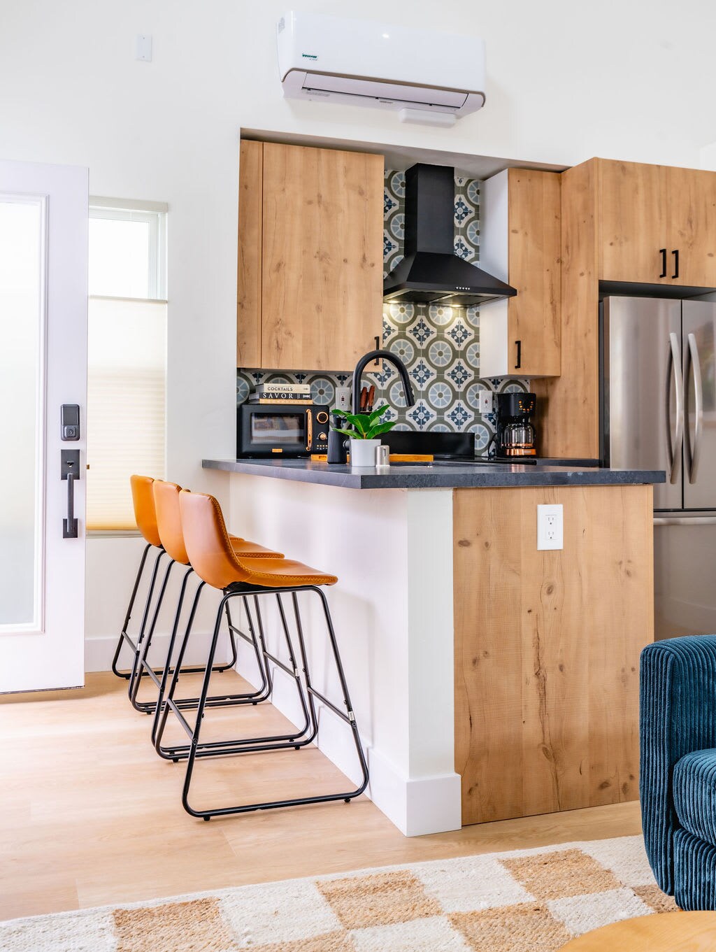 The kitchen features modern cabinetry with a stylish dark countertop. Three sleek barstools offer seating at the breakfast bar, which is complemented by decorative backsplash tiles. A stainless steel refrigerator and microwave are visible, along with a coffee maker for convenience.