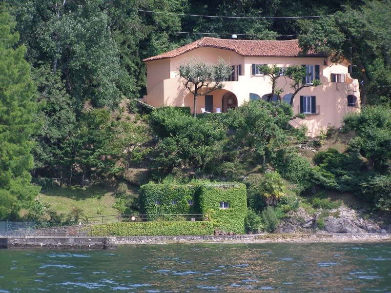 A villa is positioned on a hillside above the lake, showcasing peach-colored walls and a tiled roof. Lush greenery surrounds the property, while steps lead down to a small beach area. The building features multiple windows, some open, offering views of the lakeside.