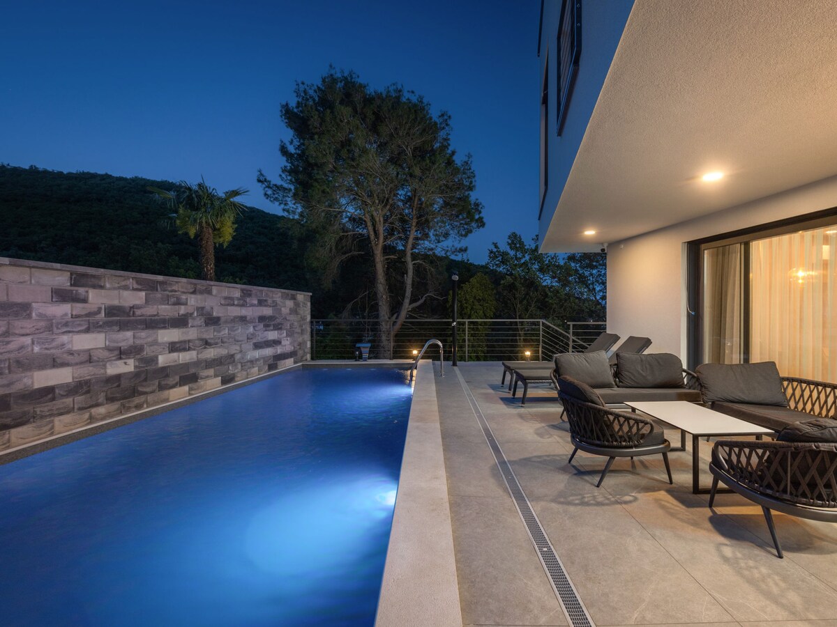 An angular swimming pool is illuminated in blue at dusk, surrounded by a smooth tiled deck. Comfortable lounge chairs are situated nearby, with a modern table positioned between them. Lush greenery is visible in the background, enhancing the serene atmosphere.