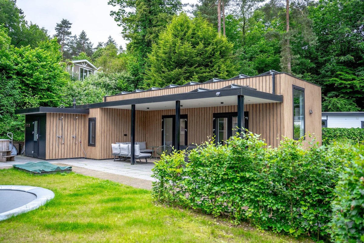 A modern wooden cottage is surrounded by lush greenery, featuring large windows and a spacious patio area furnished with outdoor seating. A trampoline is visible in the yard, complementing the serene outdoor environment.