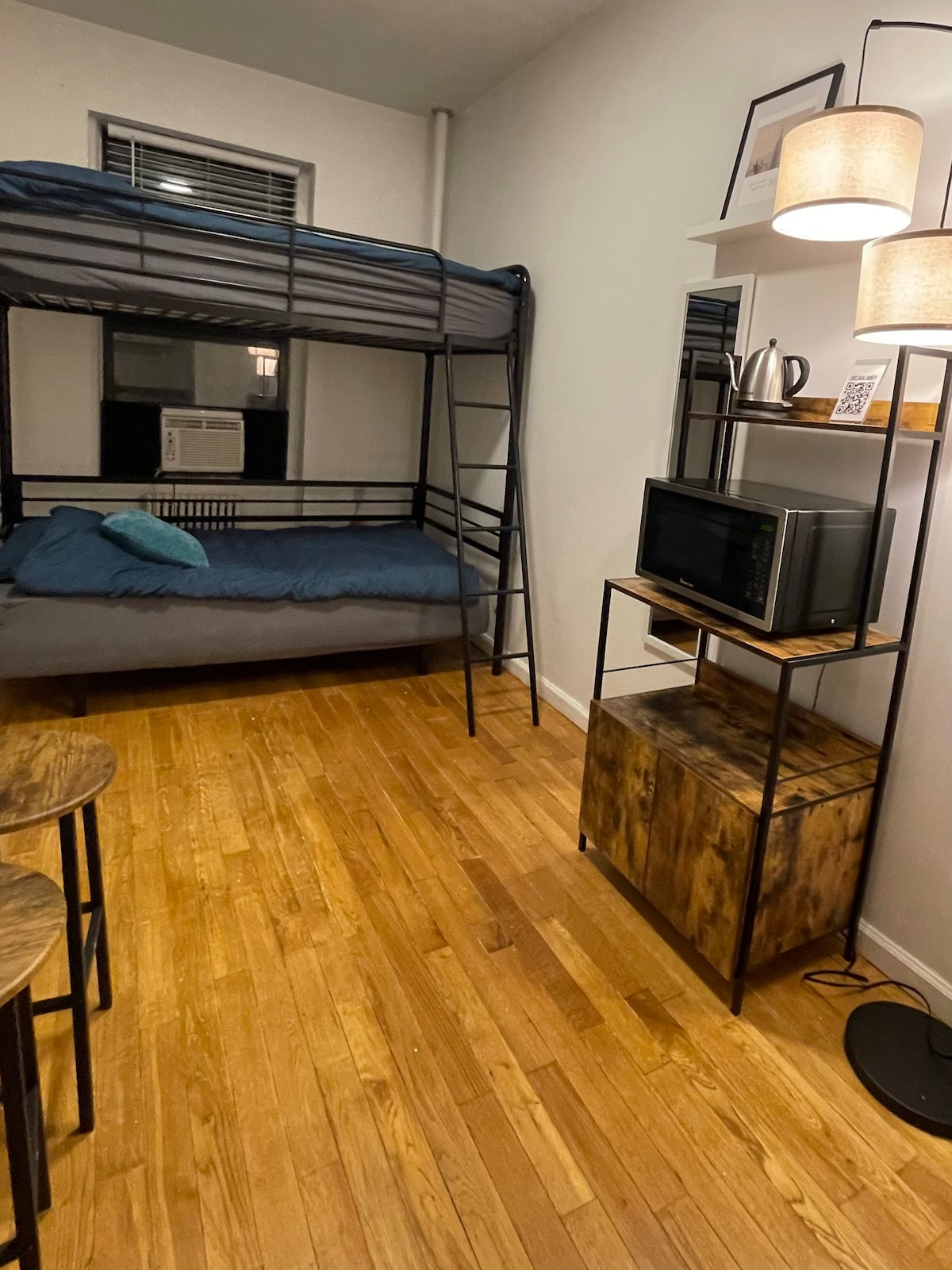 A room features a twin-over-twin bunk bed alongside a cozy sleeping area. A small kitchenette with a microwave is visible within a metal shelving unit. Wooden flooring is present, complemented by two round stools. Natural light enters through the window, enhancing the space.