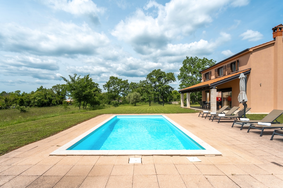 A rectangular swimming pool with clear blue water is featured in a spacious outdoor area. Surrounding the pool are several loungers and a few umbrellas, providing options for relaxation. The landscape includes lush greenery and trees, contributing to a serene atmosphere.