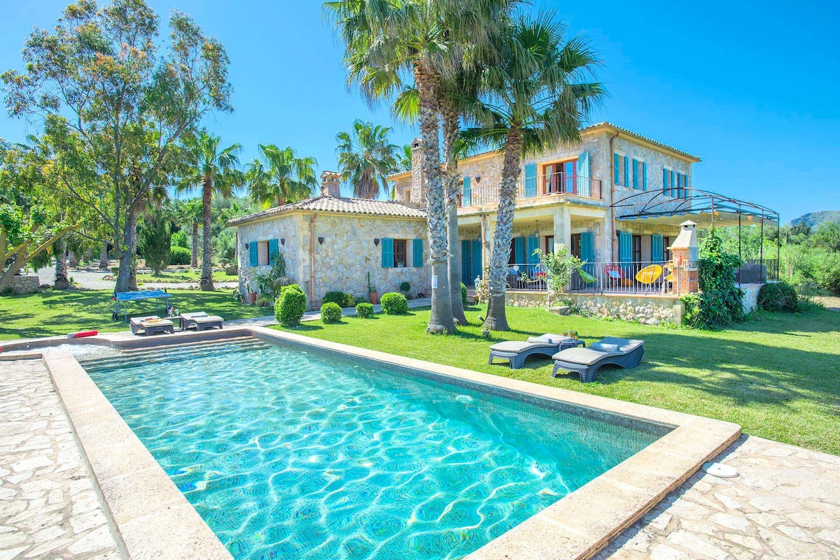 The exterior of a spacious villa is highlighted, featuring a private swimming pool surrounded by palm trees and neatly trimmed grass. Sun loungers are positioned by the pool, and the building showcases a combination of stone and vibrant colors, with several windows visible and a covered terrace area.