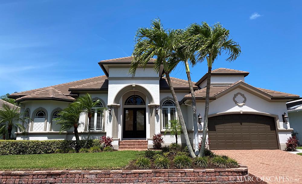 A modern villa is showcased with a symmetrical façade featuring arched entrances and a tiled roof. Lush palm trees and manicured landscaping frame the exterior, while a spacious driveway leads to a two-car garage, enhancing the welcoming appeal of this elegant home.