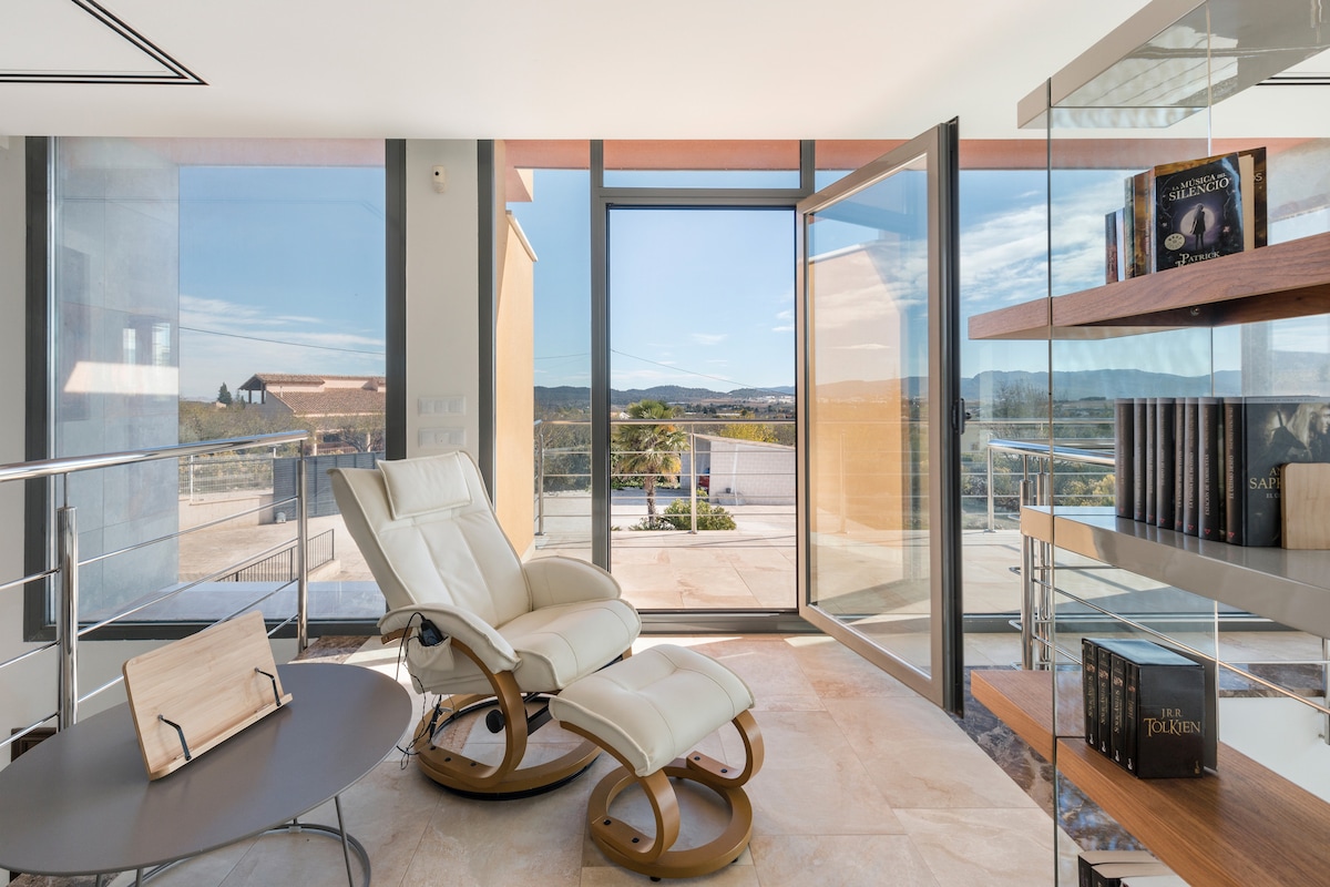A bright and airy reading nook is observed, featuring a comfortable reclining chair positioned beside a sleek table. Large glass doors open to a scenic view, allowing natural light to fill the space. Books are neatly arranged on wooden shelves nearby.