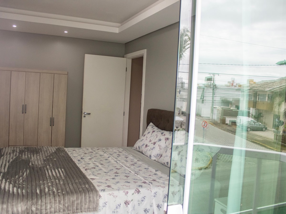 A cozy bedroom showcases a spacious bed dressed in floral linens. A large wardrobe is positioned against the wall, with soft lighting enhancing the room's ambiance. A partial view of the outside is visible through glass doors, providing natural light.