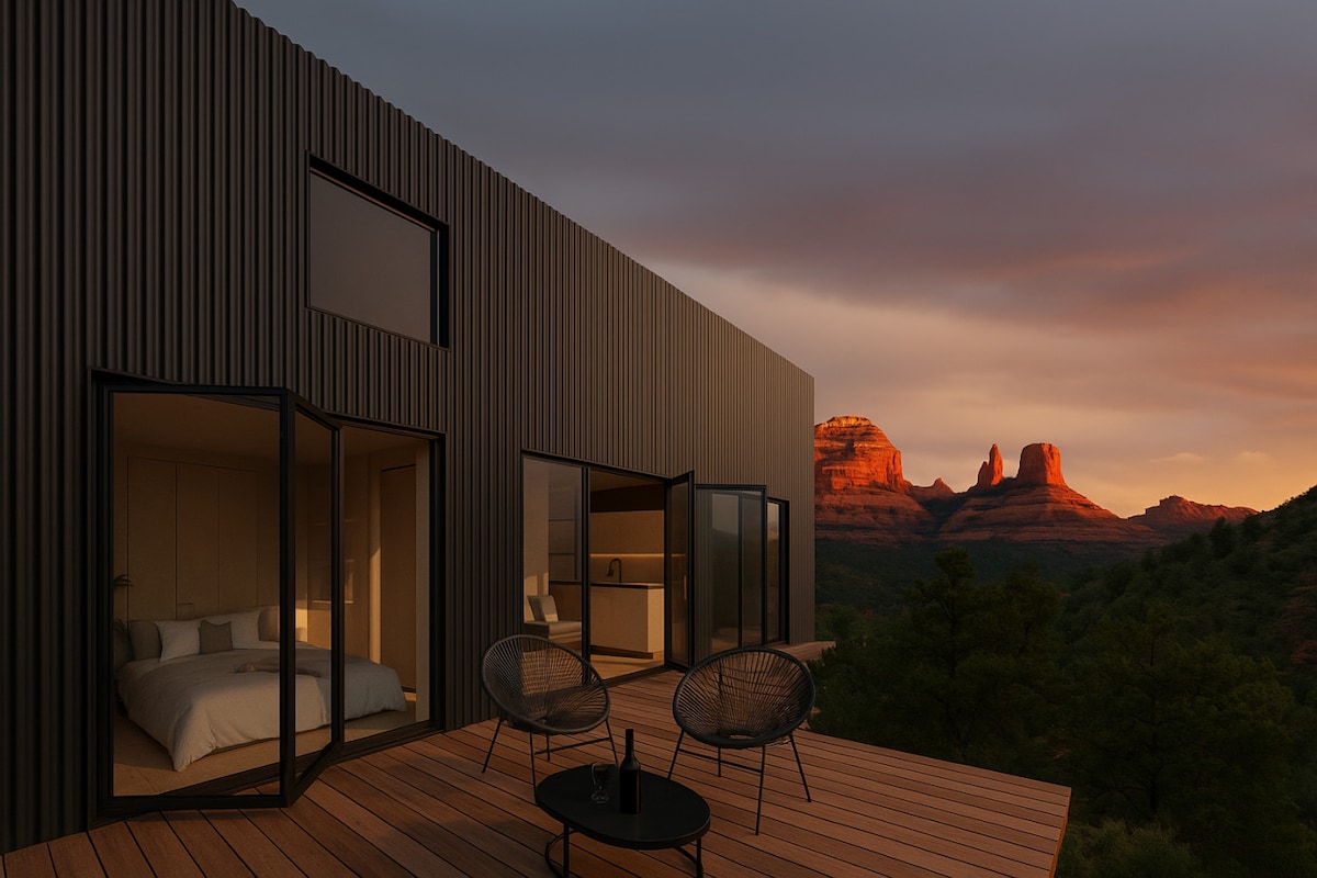 A modern cabin exterior displays expansive glass doors opening onto a wooden deck. Comfortable outdoor seating is arranged for relaxation, offering views of the surrounding canyon landscape. The warm glow of sunset highlights distant rock formations, creating a serene backdrop.
