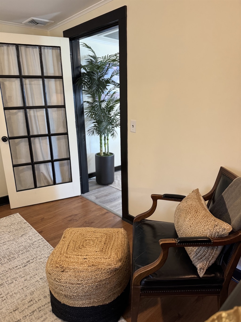 A stylish seating area is depicted, featuring an elegant chair paired with a textured pouf. A plant in a cylindrical pot adds a touch of greenery, while a partially open glass-paneled door leads to another room. Warm, natural light enhances the inviting ambiance.