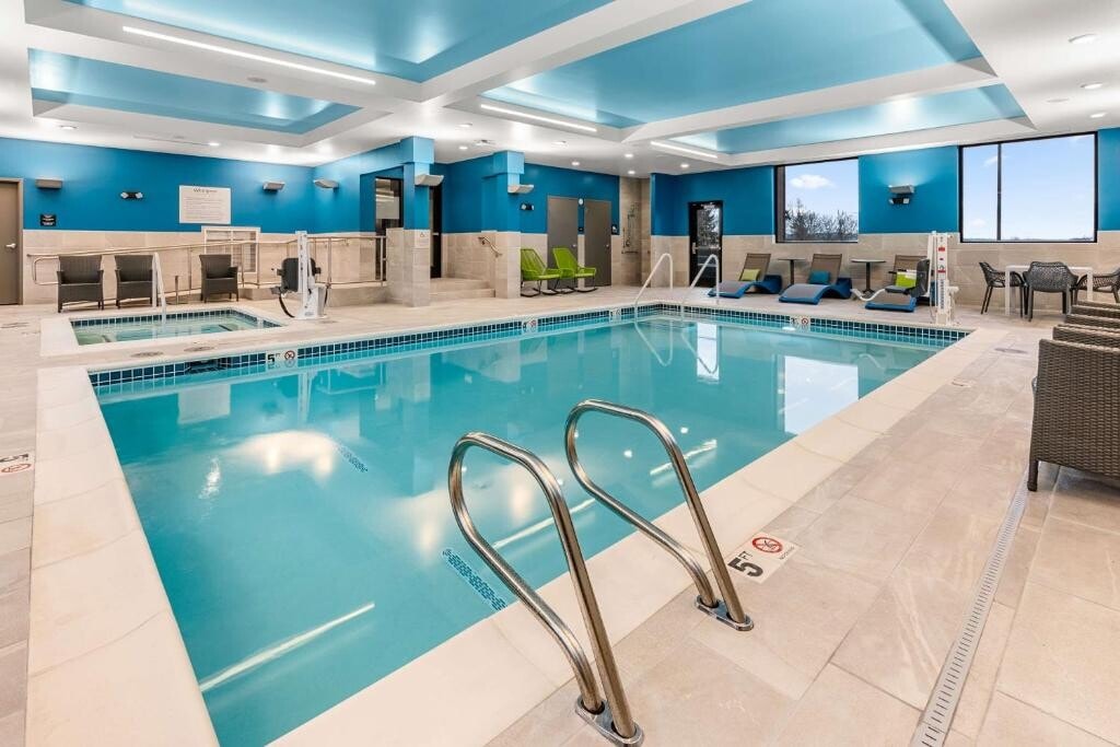An indoor swimming pool area features a clear blue water surface, surrounded by tiled flooring and light-colored walls. Lounge chairs in various colors line one side, while comfortable seating is provided in a corner. Large windows allow natural light to fill the space.