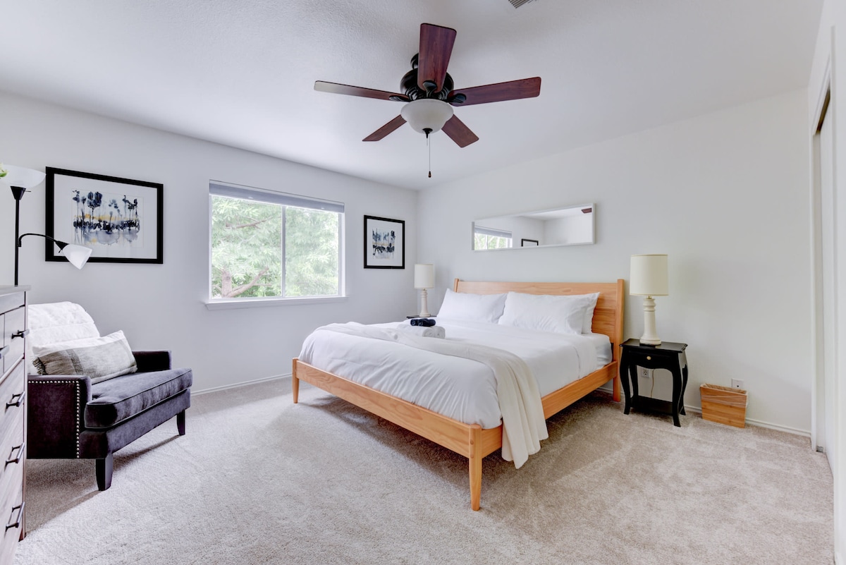 A spacious bedroom is furnished with a large wooden bed topped with crisp white linens. Soft carpeting covers the floor, and a window invites natural light. A comfortable armchair and a floor lamp are positioned beside a dresser, creating a cozy atmosphere for relaxation.