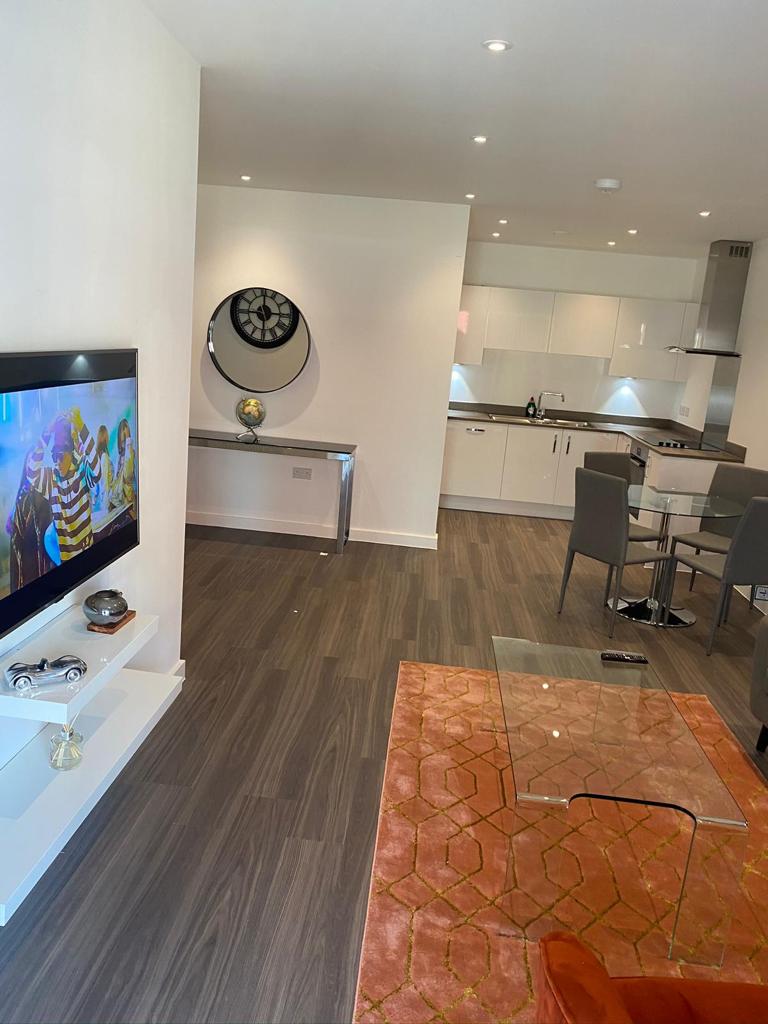 An open-concept living area is featured, showcasing a sleek television mounted on the wall. A round wall mirror reflects light, while a modern glass dining table is surrounded by grey chairs. A vibrant orange rug adds warmth to the space, complementing the neutral color palette.
