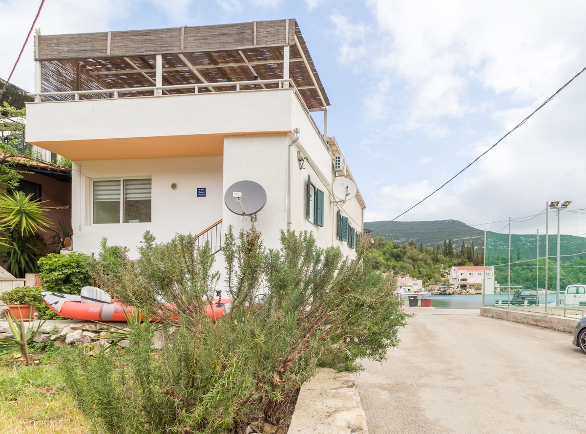 The exterior of a two-story holiday home is visible, featuring a balcony with wooden decking. Surrounding greenery adds a natural touch, while a satellite dish is mounted on the wall. A paved road leads to the property, with views of nearby hills and a small waterway.