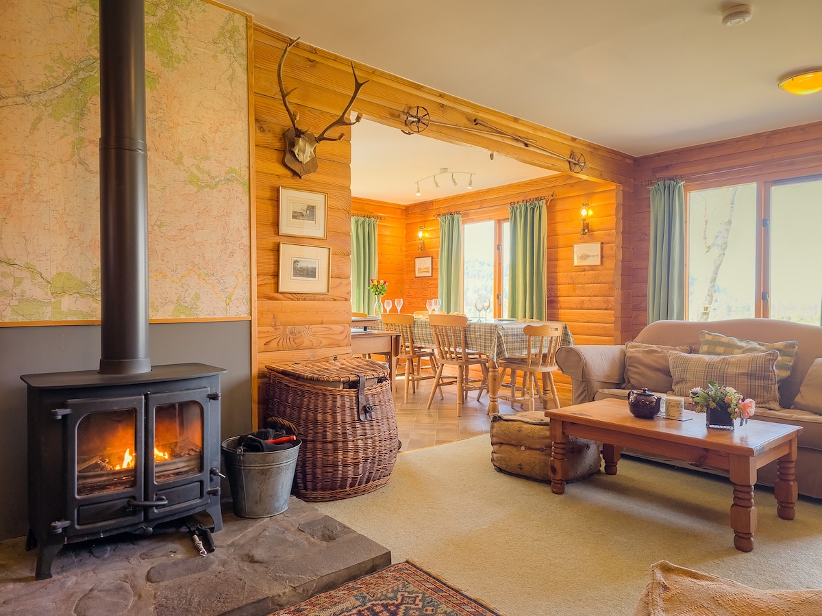 A welcoming living area features a wood-burning stove, providing warmth beside a rustic stone hearth. A large map adorns one wall, complemented by wooden accents. Large windows offer calming views, while a dining table is set, ready to accommodate guests. Comfortable seating invites relaxation.