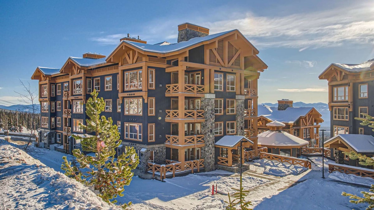 The condominium building is designed with a rustic mountain aesthetic, featuring dark wood siding and large balconies. Snow covers the surrounding landscape, and the building is set against a backdrop of mountains under a clear blue sky, creating an inviting winter atmosphere.