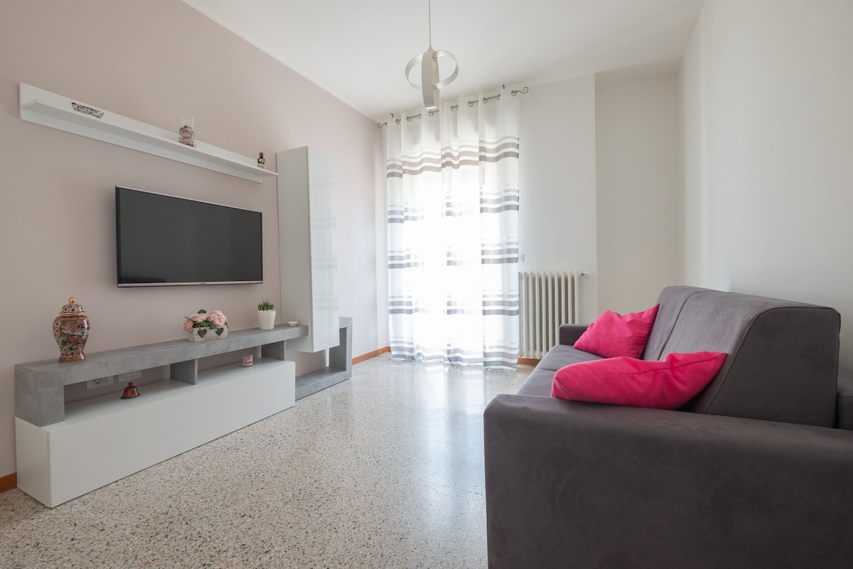 The living room showcases a neutral-toned sofa with bright pink cushions, positioned opposite a wall-mounted television. A low, modern console table holds decorative items. Natural light filters through sheer curtains, illuminating the space and highlighting the polished floor.
