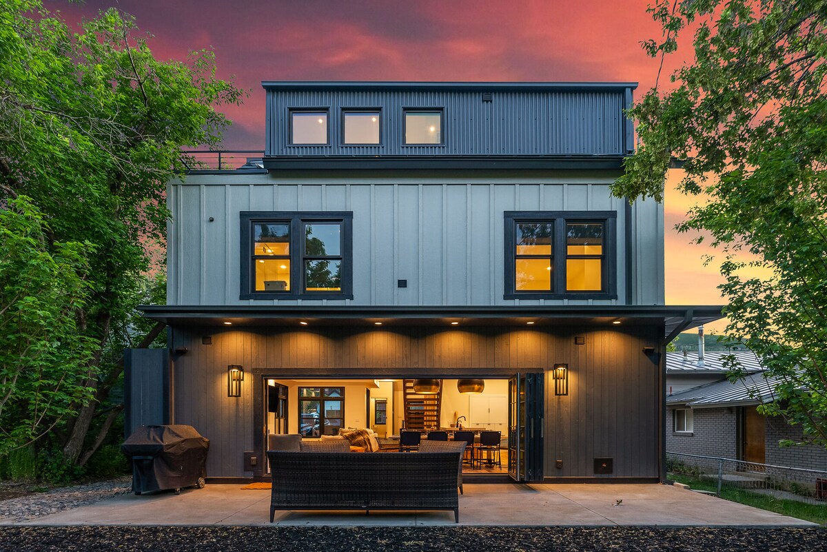 The exterior of the vacation home is showcased against a vibrant sunset. The structure features a mix of horizontal and vertical elements, large windows, and a spacious patio area furnished with comfortable seating. Surrounding greenery adds to the serene atmosphere.