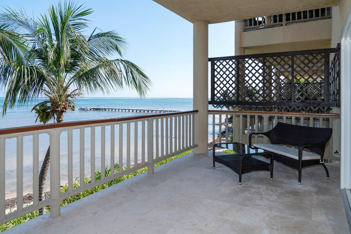 A private balcony provides a comfortable seating area with rattan chairs, overlooking the shimmering waters of the Caribbean Sea. Lush palm trees sway gently in the breeze, framing views of a nearby pier and the serene horizon.