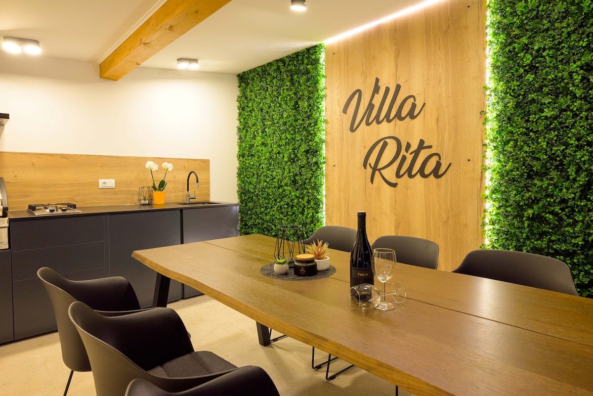 A contemporary dining area features a large wooden table surrounded by black chairs, set against a backdrop of greenery and wooden accents. The space is illuminated by warm lighting, with a kitchen area visible in the background, providing a welcoming environment for meals.