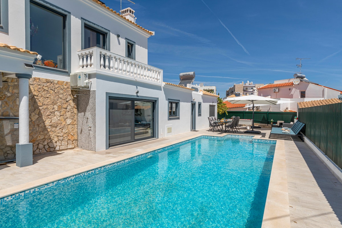 A serene outdoor space features a refreshing swimming pool surrounded by a sunbathing area with lounge chairs. The villa, showcasing a blend of stone and smooth surfaces, showcases floor-to-ceiling glass doors that invite natural light into the adjoining living area.