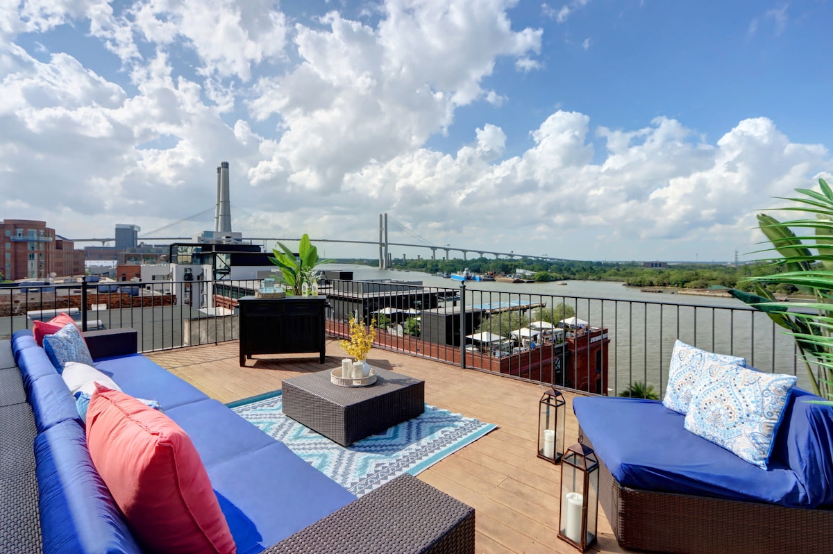 A rooftop terrace is presented, featuring comfortable seating with blue cushions and pink accent pillows. A low coffee table is centered on a colorful rug, with decorative lanterns and a vase of flowers. The Savannah River and a bridge can be seen in the background under a partly cloudy sky.