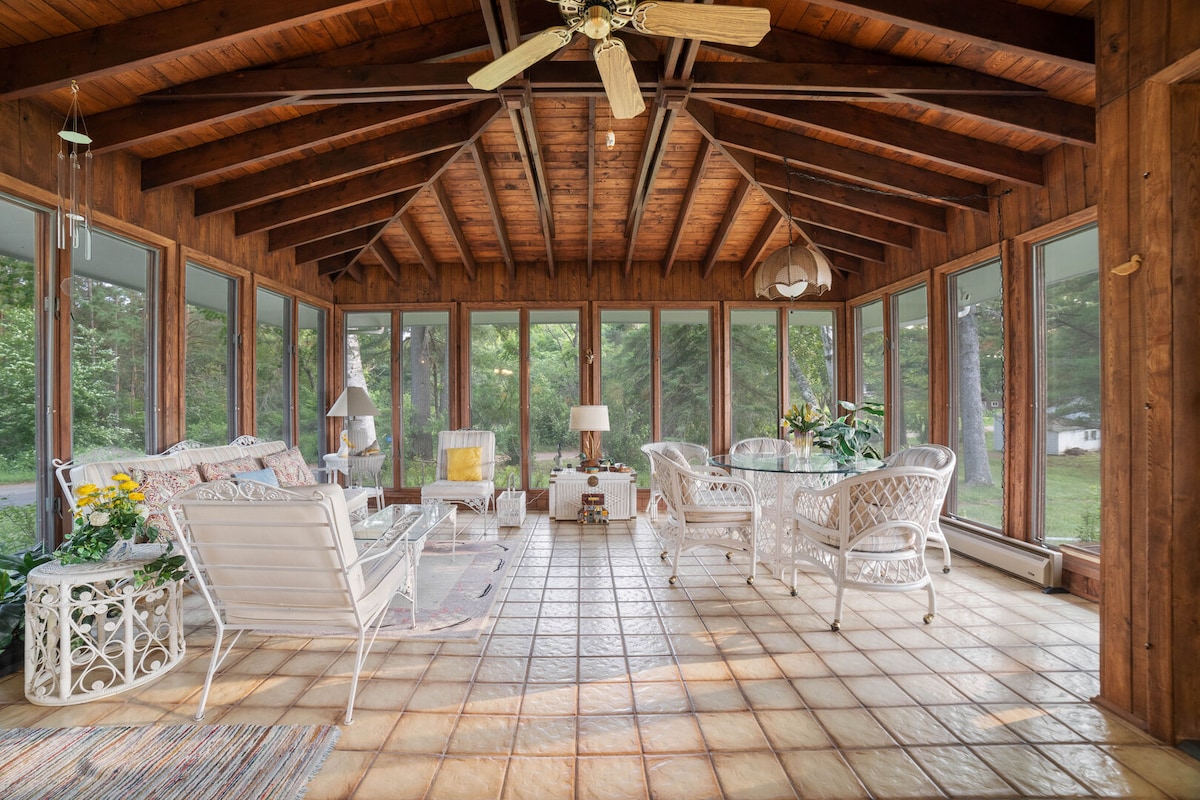 A sunroom features large windows allowing natural light to fill the space. A wooden ceiling with exposed beams enhances the rustic charm. Seating includes white wicker furniture, complemented by floral accents and a small side table. A dining area is visible, providing a cozy setting for meals.