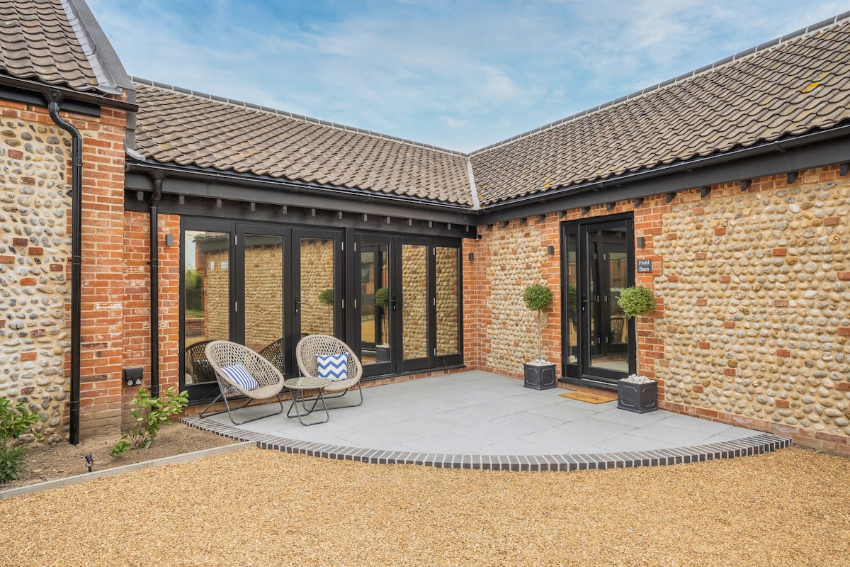 A charming outdoor seating area features two stylish chairs positioned on a paved surface. Stone walls and a gravel pathway create a welcoming atmosphere. Floor-to-ceiling glass doors reflect natural light, showcasing the connection between the interior and exterior spaces.