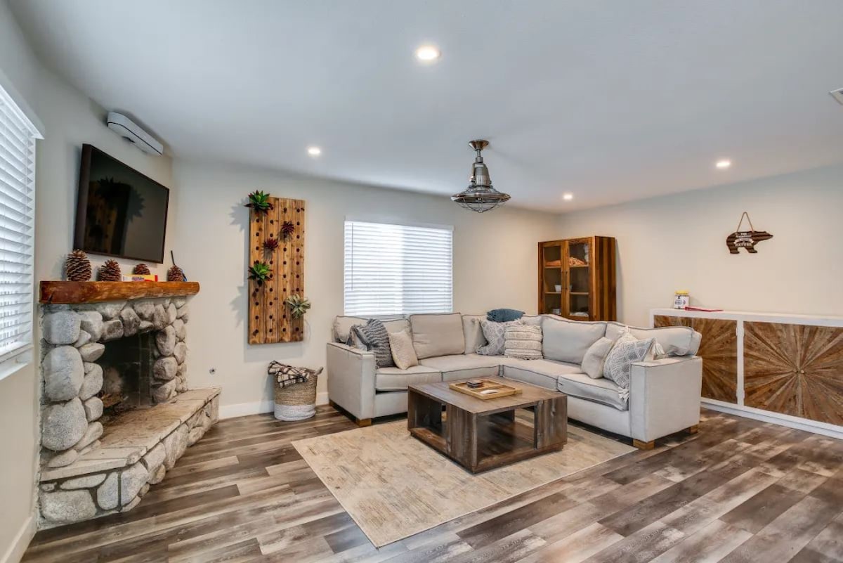 A spacious living area features a comfortable sectional sofa arranged around a wooden coffee table. A stone fireplace serves as a focal point, complemented by natural decor on the walls. A large window provides ample light, enhancing the inviting ambiance.