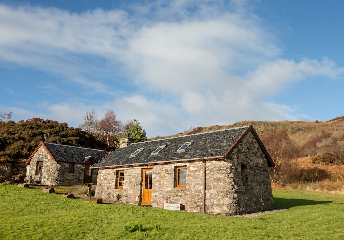 The exterior of the main cottage is presented, featuring robust stone walls and a slate roof. Skylights enhance natural daylight within. The cottage is set amidst a lush green lawn, bordered by gentle hills, with an adjacent byre visible in the background.
