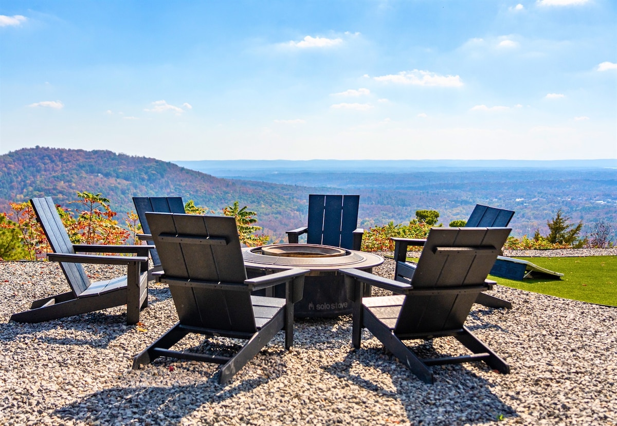 A circular arrangement of Adirondack chairs encircles a central fire pit, set against a backdrop of expansive mountain vistas. The area is surrounded by colorful foliage, and the blue sky stretches overhead, creating a serene outdoor space for gatherings.