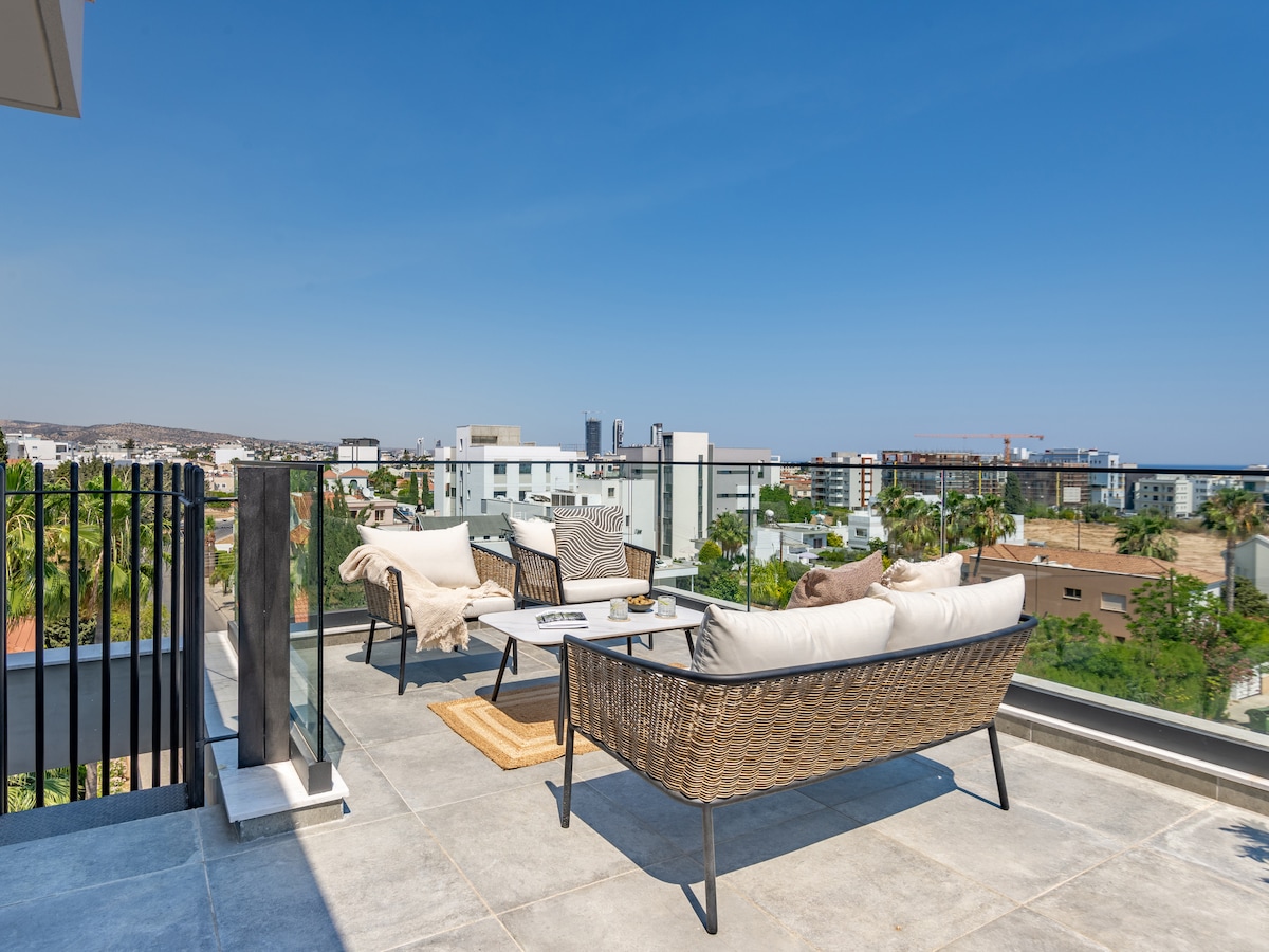 A rooftop terrace is presented, featuring comfortable seating arrangements on wicker chairs and a cozy loveseat. A small table holds decorative items, while a view of the surrounding cityscape and greenery is visible under clear blue skies.