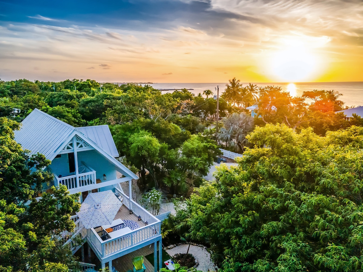 A view of the welcoming home surrounded by lush tropical greenery, with a balcony showcasing outdoor seating. The sun sets over the water, casting a warm glow across the horizon, while palm trees gently sway in the breeze, enhancing the serene landscape.