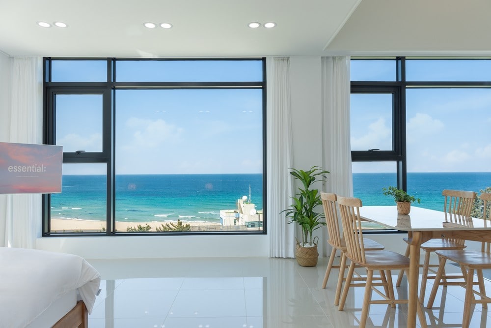 A bright, airy room is characterized by large windows offering uninterrupted views of the ocean and coastline. A dining table surrounded by wooden chairs is positioned beside the windows, while a spacious bed is visible in the foreground, enhancing the tranquil atmosphere.