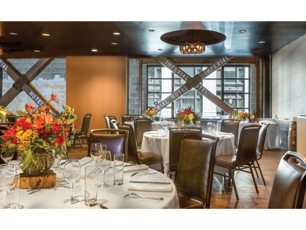 An elegantly arranged dining space features round tables adorned with white tablecloths and vibrant floral centerpieces. Leather chairs surround the tables, and large windows offer ample natural light filtered through decorative accents. A warm tone is created by the combination of dark walls and lighting fixtures.